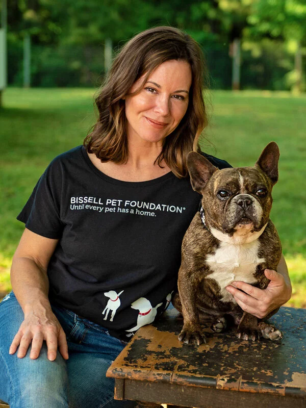 A woman with shoulder-length brown hair sits outdoors on a bench, smiling and holding a brindle French Bulldog. She wears a black Bissell Pet Foundation t-shirt. Green grass and trees are in the background.
