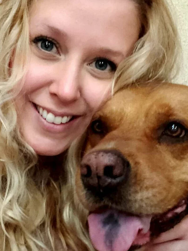 A woman with long, wavy blonde hair smiles beside a brown dog with its tongue out, both looking at the camera in a close-up selfie.