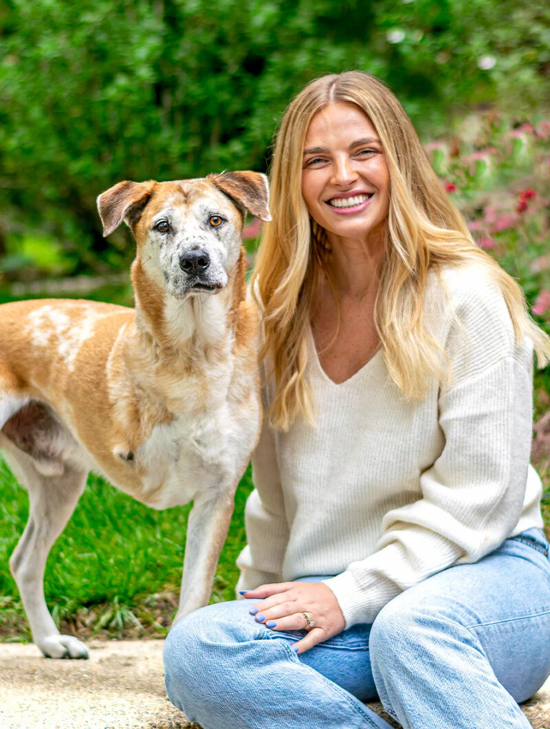 A smiling woman with long blonde hair and a light sweater sits outdoors on the grass next to a brown and white dog with one ear up, both looking at the camera.
