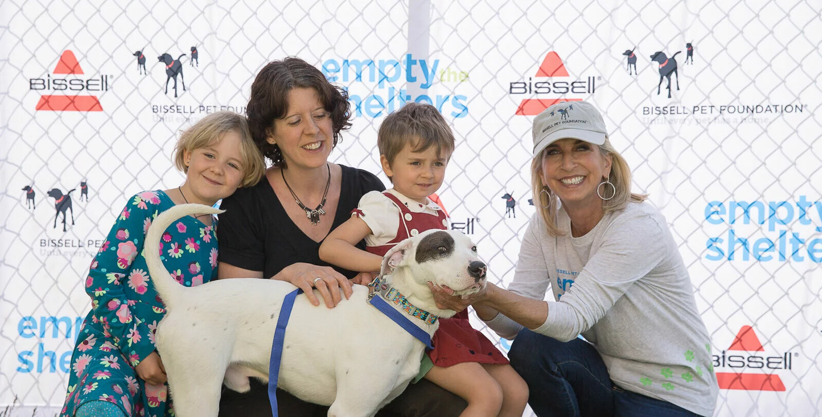 Two women and two children smile while posing with a white dog with brown spots in front of a fence and a backdrop reading “empty the shelters,” supporting the Bissell Pet Foundation.