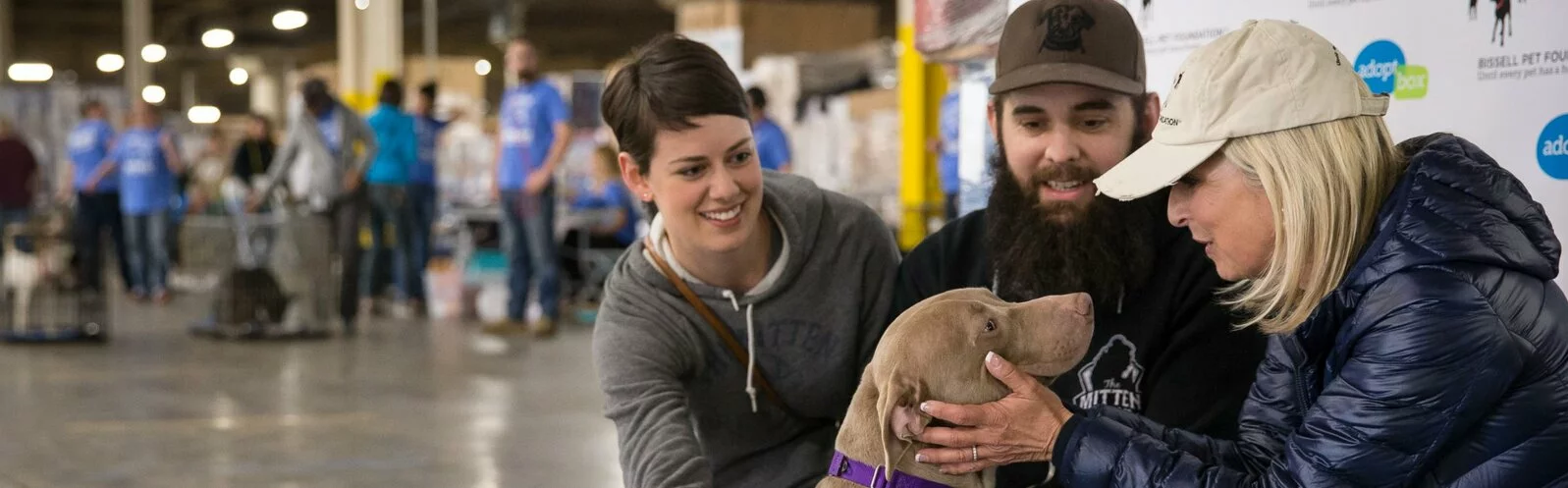 Three people sit indoors at an adoption event, smiling and interacting with a light brown dog wearing a purple collar. Other people and dogs are visible in the background of the large space.