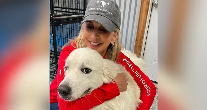 A smiling woman in a Bissell Pet Foundation hat and red shirt hugs a large, fluffy white dog in a Bissell Pet Foundation bandana, inside a room with a kennel and white door—highlighting hope amid the ongoing Shelter Crisis.