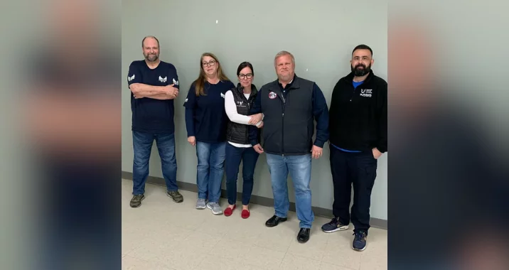 Five adults stand side by side against a gray wall indoors, casually dressed in jeans and jackets, smiling at the camera. The informal setting suggests they may have just finished a training session together.