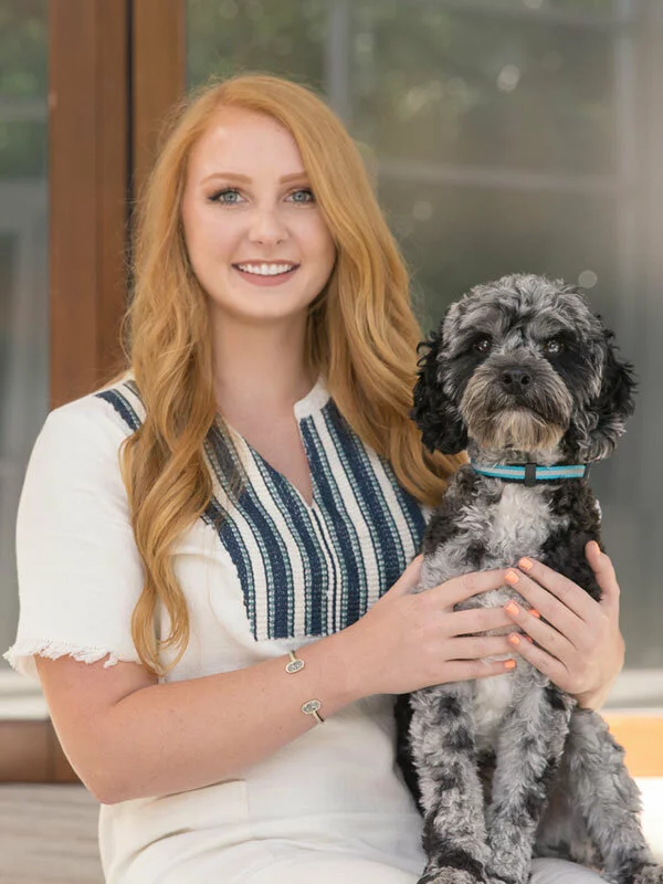 A woman with long red hair wearing a white dress with blue embroidery sits and smiles, holding a black and gray dog with a blue collar on her lap.
