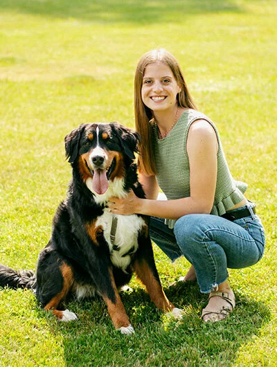 A woman with long light brown hair, wearing a sleeveless top and jeans, kneels on grass next to a large, happy Bernese Mountain Dog. Both are looking at the camera and smiling.
