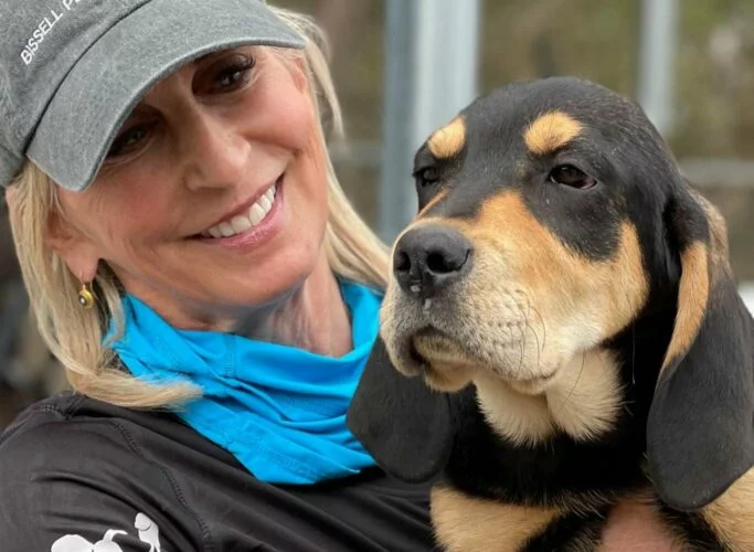 A smiling woman wearing a gray cap and blue scarf holds a large black and tan dog, both looking content and happy outdoors.