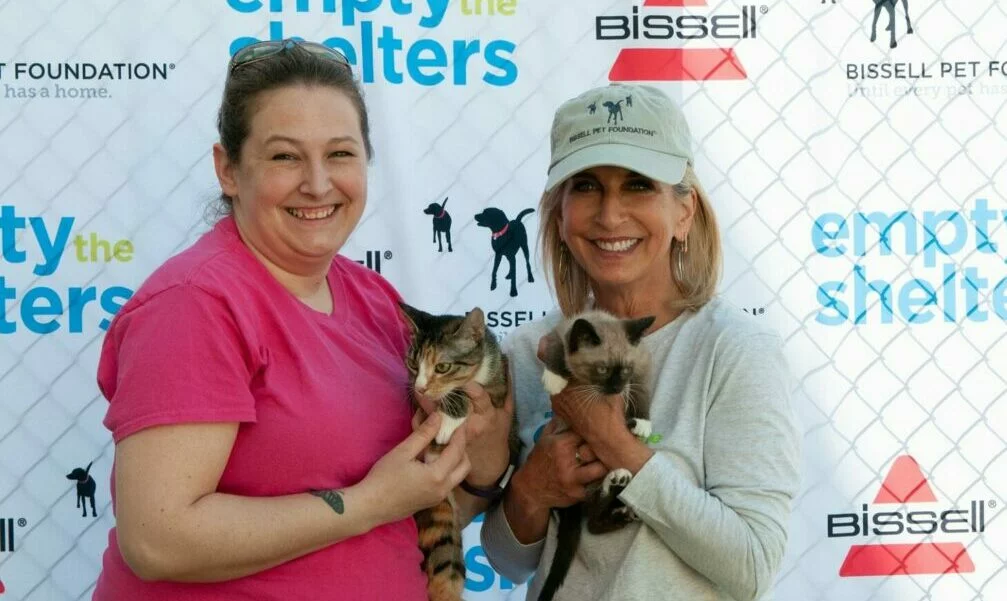 Two women smile while holding cats in front of a fence with an “Empty the Shelters” and “Bissell Pet Foundation” banner in the background. One woman wears a pink shirt; the other wears a light hat and shirt.