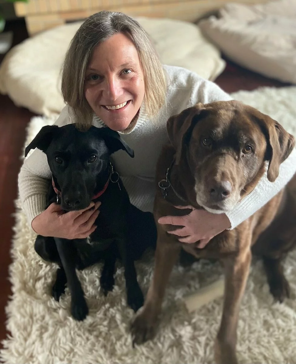 A smiling person with short light hair sits on a fluffy rug, hugging a black dog and a brown dog, both of whom are looking at the camera. Cozy beds are visible in the background.