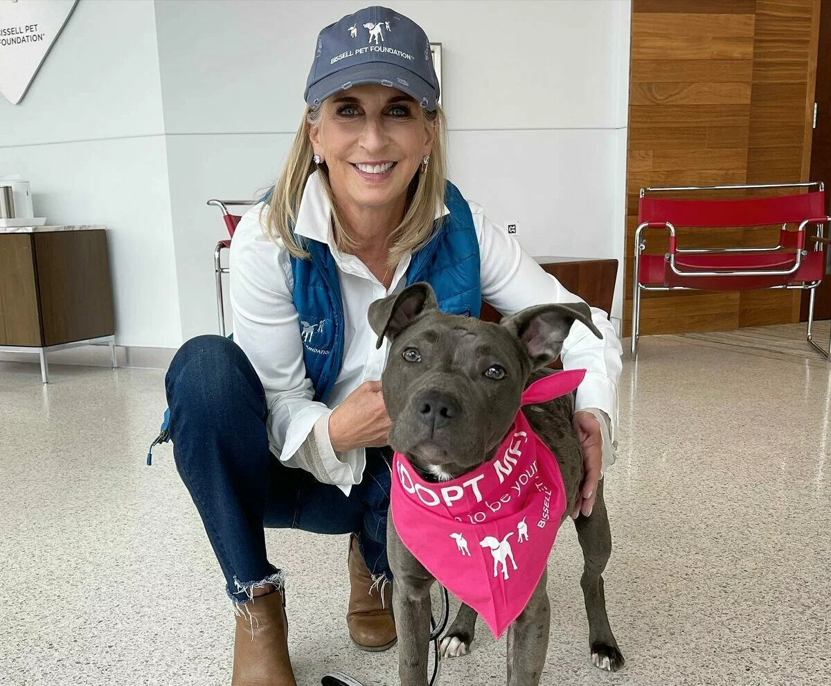 A smiling woman in a blue cap and vest crouches next to a gray dog wearing a pink Adopt Me bandana inside a modern building with red chairs and wooden accents.
