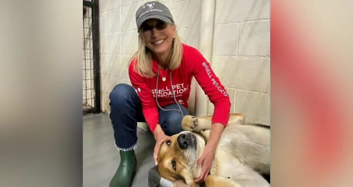 A smiling woman in a red shirt and cap kneels beside a large dog lying on the floor, gently petting its head. The dog looks relaxed and content inside what appears to be an animal shelter.