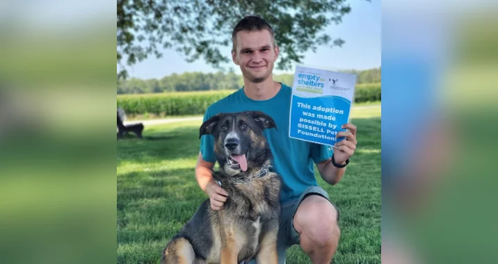 A smiling man kneels on grass next to a German Shepherd mix dog. He holds a blue sign that reads, “This adoption was made possible by BISSELL Pet Foundation.” Trees and fields are in the background.