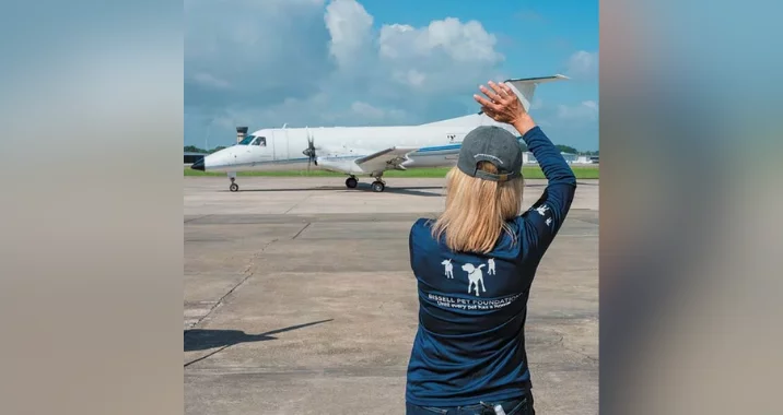 A woman with blonde hair waves at a small white airplane on a runway under a blue sky with clouds. She is wearing a navy shirt with graphics and text on the back.
