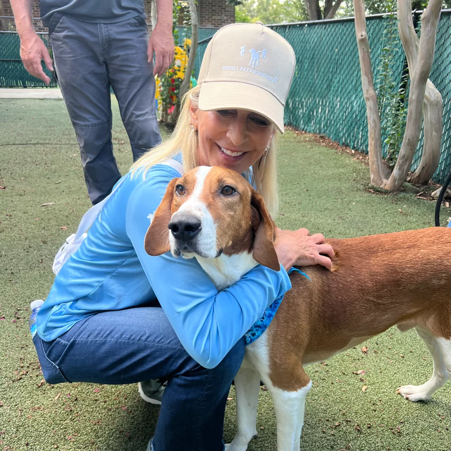 A smiling woman in a light blue shirt and tan cap hugs a brown and white dog outdoors on green turf, with a person standing in the background near a green chain-link fence and leafy trees.