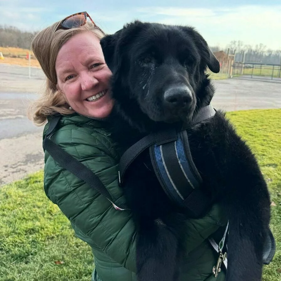 A smiling woman in a green jacket hugs a large, fluffy black dog outdoors on a grassy area, with a fence and trees visible in the background.