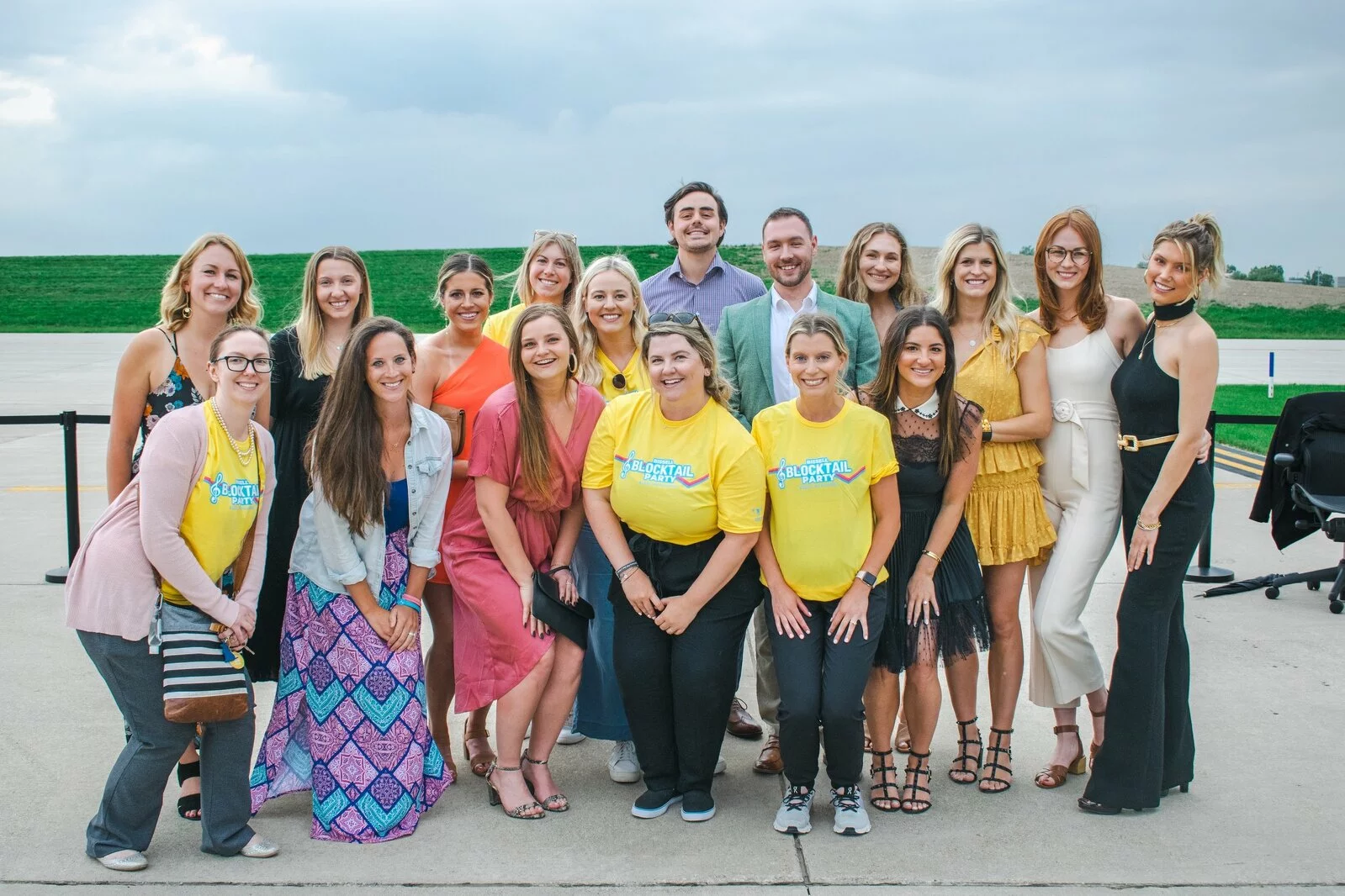 A group of 18 people, including men and women, pose and smile outdoors on a paved area. Three women in front wear bright yellow shirts, while others wear casual or dressy clothing. A grassy field and cloudy sky are in the background.