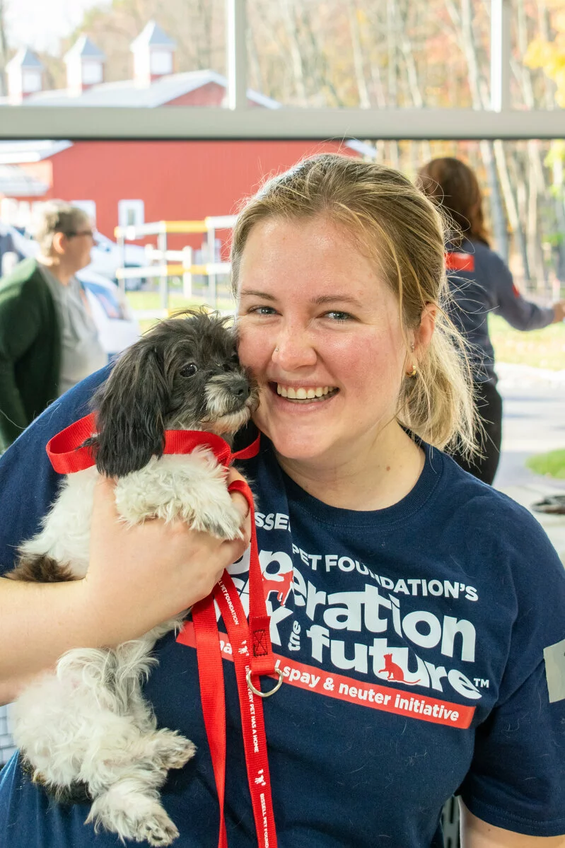 A smiling woman wearing a navy blue T-shirt holds a small black and white dog with a red ribbon around its neck. They are indoors with people and a red barn visible through the window behind them.