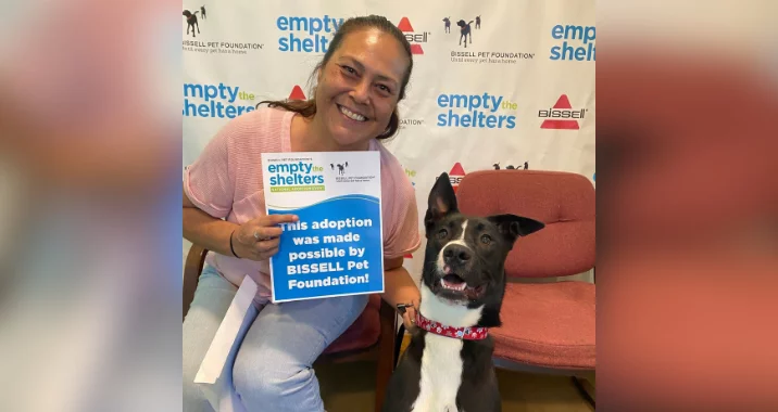 A smiling woman sits beside a happy black and white dog, holding a sign that reads, This adoption was made possible by BISSELL Pet Foundation! The background features Empty the Shelters and BISSELL logos.