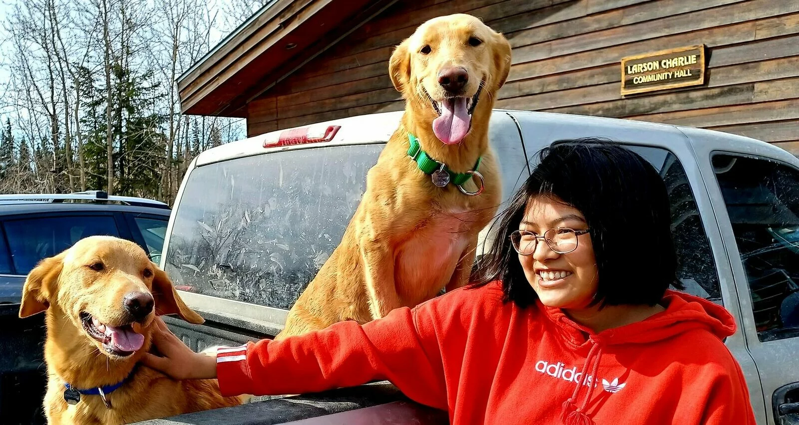 A smiling person in a red Adidas hoodie stands by a pickup truck, petting two happy golden dogs in the truck bed, with a wooden building and trees in the background.