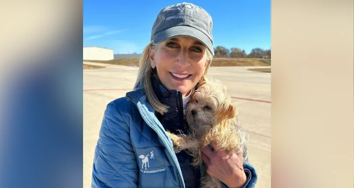 A smiling woman in a gray cap and blue jacket holds a small, scruffy light-brown dog outdoors on a sunny day, with a clear sky and buildings visible in the background.