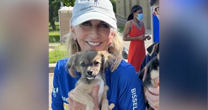 A smiling woman in a blue shirt and gray cap holds a small brown and white puppy outdoors. People and another puppy are visible in the background, including a woman in a red dress and a blue face mask.