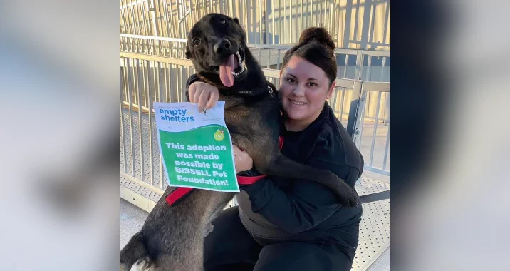 A woman in black clothing smiles while hugging a large, happy black dog. The dog holds a sign that reads, This adoption was made possible by BISSELL Pet Foundation! They are seated on a metal bench outdoors.