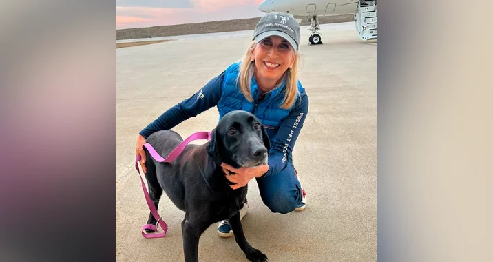 A woman wearing a blue vest and cap smiles while kneeling next to a black dog with a pink leash on an airport tarmac, with a small jet in the background at sunset.