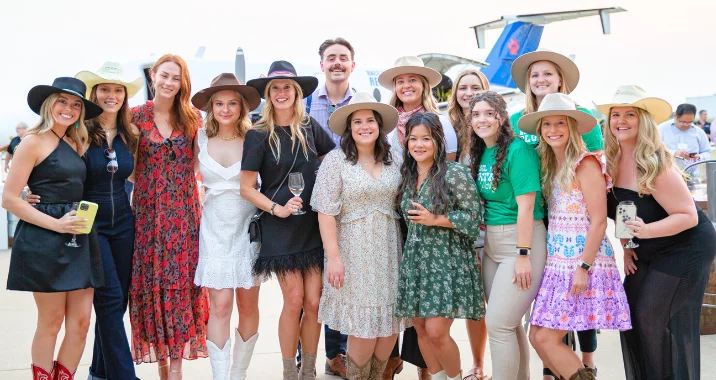 A group of people, mostly women in dresses and hats, pose together outdoors. Many are smiling and holding drinks, and there is a blue helicopter visible in the background.