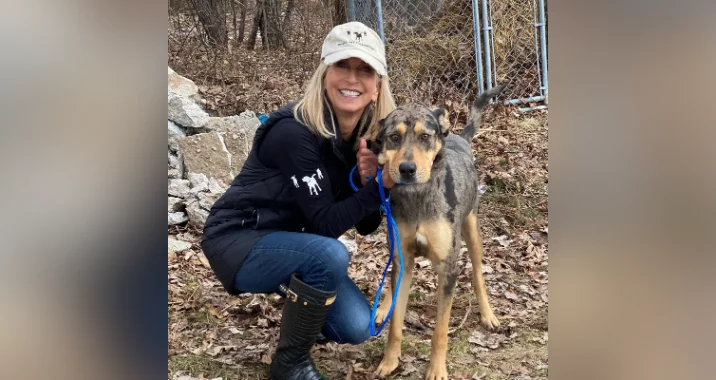 A smiling woman in outdoor clothing and a hat kneels beside a large dog on a blue leash. They are outside on fallen leaves near a chain-link fence and some trees.