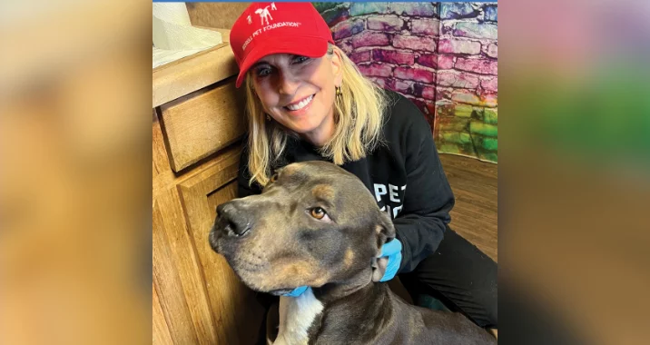 A smiling woman in a red cap and black shirt kneels indoors beside a large gray and white dog, gently holding its head. The background features a colorful brick wall.