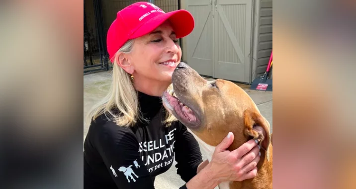 A smiling woman wearing a red cap and black shirt hugs a brown and white dog, who is nuzzling her chin affectionately outdoors near a shed.