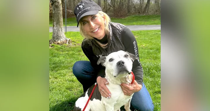 A woman in a black cap and jacket kneels on grass, smiling and hugging a white and brown dog on a red leash. Trees and a path are visible in the background.