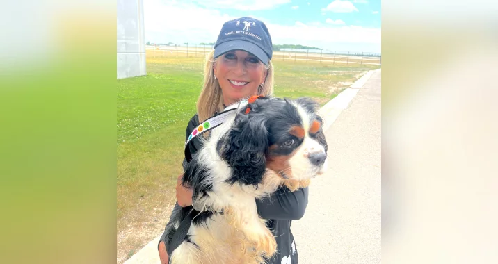 A smiling woman wearing a dark baseball cap and sunglasses holds a large, fluffy tricolor dog outside on a sunny day near a grassy area and a pathway.