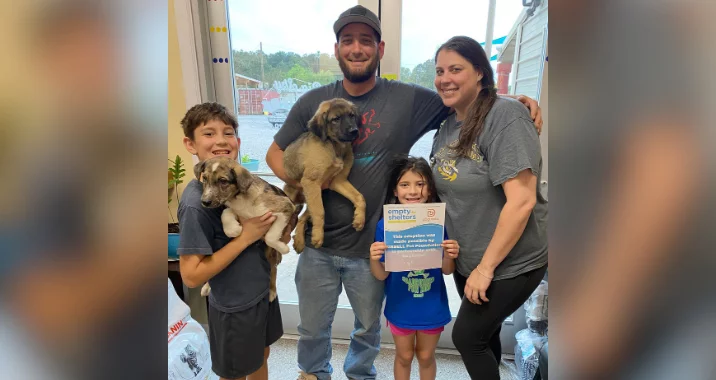 A smiling family of four stands indoors holding two puppies. The father and son each hold a puppy. The young daughter holds a certificate and stands next to her mother. They appear happy and are posing for a photo.