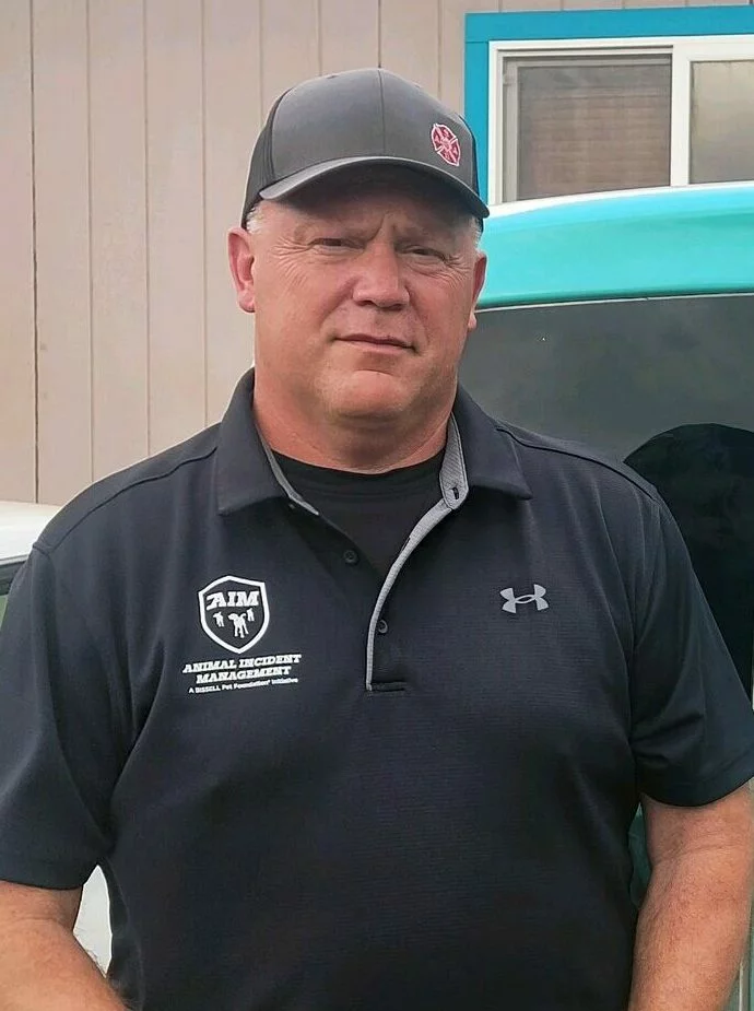 A man wearing a black baseball cap and a dark polo shirt with logos stands in front of a blue-green vehicle and a beige building. He is facing the camera with a neutral expression.