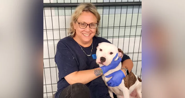 A smiling woman with glasses and blue gloves sits beside a white and brown dog, gently holding its face, in front of a metal fence.
