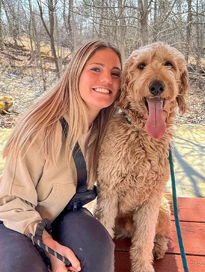 A smiling woman with long blonde hair sits next to a happy, fluffy dog with its tongue out. They are outdoors on a wooden surface with bare trees in the background.