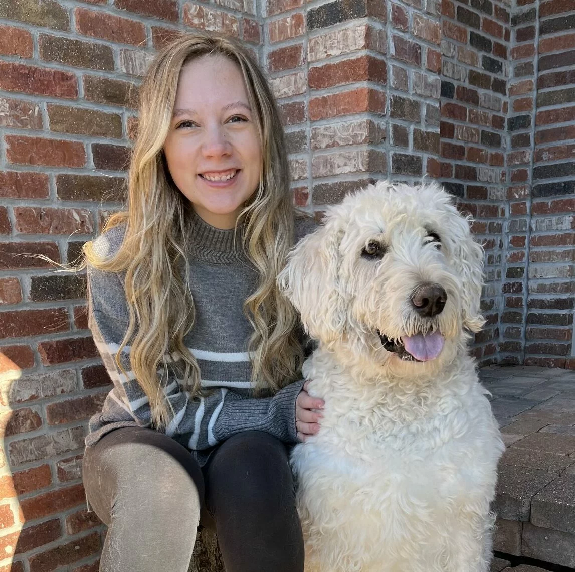 A young woman with long blonde hair and a gray sweater sits outside next to a large, fluffy white dog. They are both smiling in front of a brick wall.