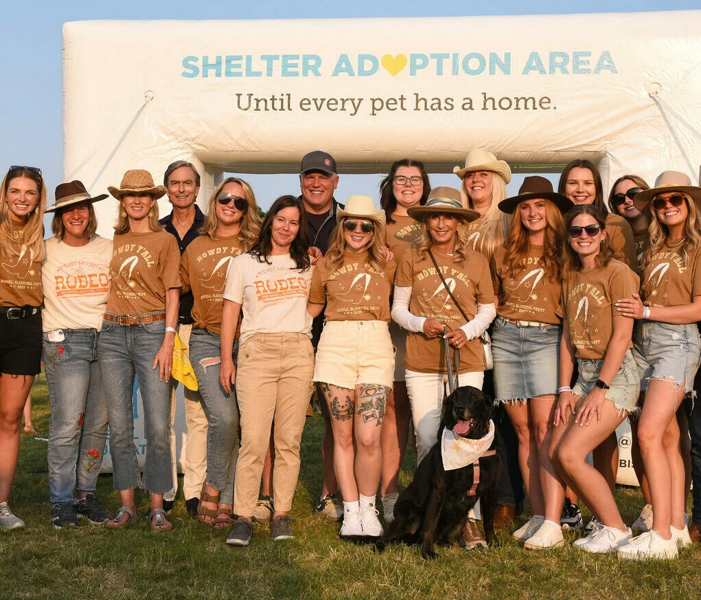 A group of people in casual clothing and hats smile together in front of a Shelter Adoption Area sign, with a black dog sitting in front. The sign reads, Until every pet has a home.