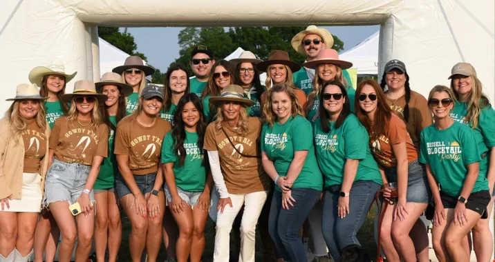 A group of people pose and smile together outdoors under an arch. Most are wearing wide-brimmed hats and casual clothes, with matching green and brown t-shirts that read “Russell Blocktall Cowgirl Camp.”.