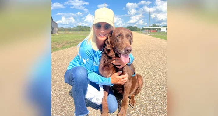 A woman in a blue shirt, jeans, and a yellow cap kneels on a path, smiling and hugging a brown dog with its tongue out. They are outdoors on a sunny day with a blue sky and clouds in the background.