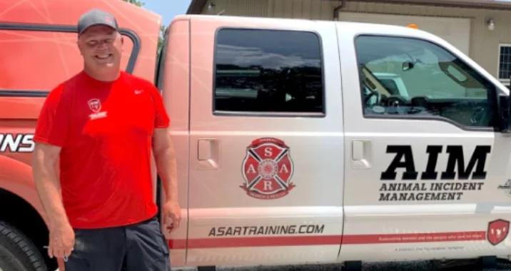A smiling man in a red T-shirt and cap stands next to a white truck labeled AIM Animal Incident Management and ASAR Training. The truck has a logo and is parked outside a building.