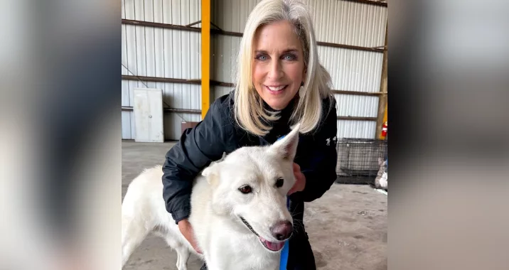 A smiling woman with blond hair poses indoors with a white dog, holding its shoulders. They appear to be in a large warehouse or industrial building with a metal wall and exposed beams in the background.