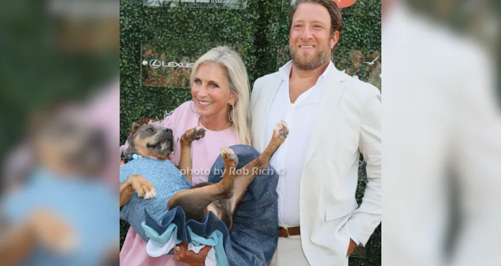 A smiling woman in a pink dress holds a relaxed dog in a blue outfit, while a man in a light suit stands beside them, all posing in front of a green leafy backdrop.