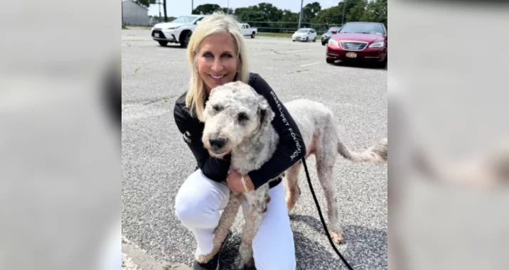A smiling woman with blonde hair crouches and hugs a large, curly-haired dog on a leash in a parking lot, with several cars and trees in the background.