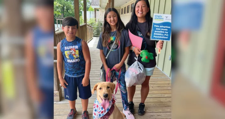 A smiling woman and two kids stand on a porch with a happy yellow dog wearing a bandana. The woman holds a sign that reads, “This adoption was made possible by BISSELL Pet Foundation.”.