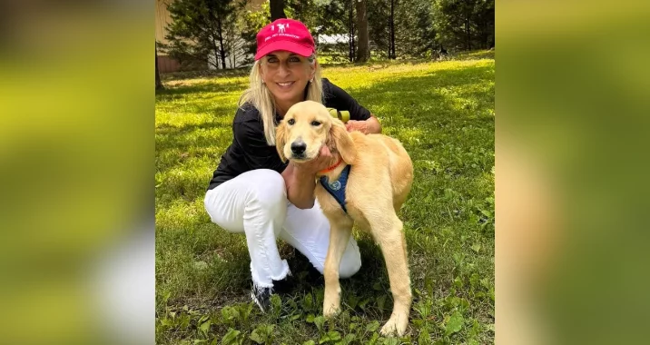A woman in a red cap, black top, and white pants kneels on the grass, smiling and hugging a golden retriever puppy wearing a harness. They are outdoors with trees and sunlight in the background.