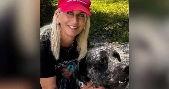 A smiling woman with long blonde hair wearing a red cap and black shirt kneels outdoors beside a large, black and white spotted dog. They are on a gravel path with green grass behind them.