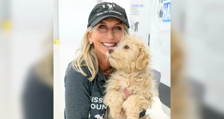 A smiling woman wearing a black cap and a grey shirt holds a fluffy, light-brown dog. The dog is nuzzling her face affectionately. The background is bright and slightly blurred.