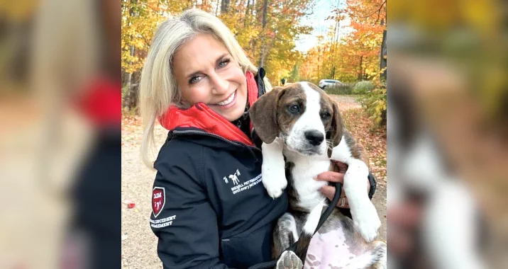 A smiling woman with blonde hair, wearing a dark jacket with red lining, holds a brown and white puppy. They are outdoors on a path lined with autumn trees, with colorful leaves in the background.