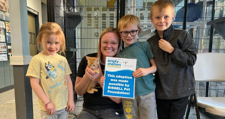 A smiling woman holding an orange kitten sits with three young children in an animal shelter. She holds a sign that reads, This adoption was made possible by BISSELL Pet Foundation! Cages and shelter posters are visible in the background.